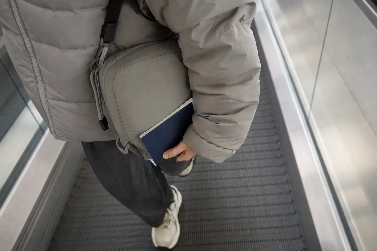 A candid-style photo of a compact crossbody bag and passport in a traveler's hand on a moving walkway; include matte nyl